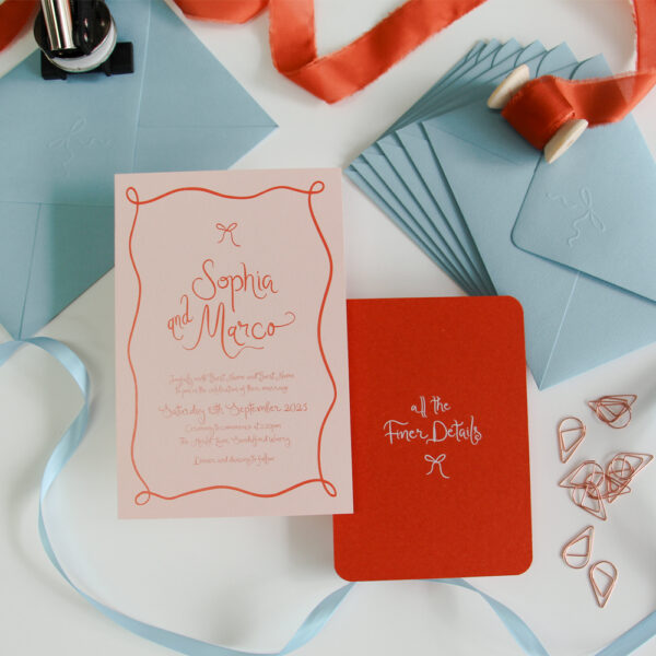 A Wedding Invitation suite including an invitation with blush pink background and orange printing. The design uses organic flowing fonts and hand drawn bows and boarders. There are blue envelopes, satin ribbon, and an envelope embosser in the background.