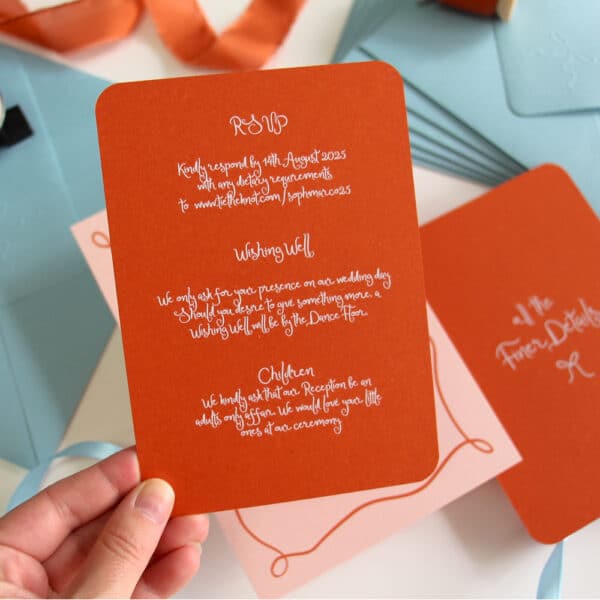A hand holding a dark orange Wedding Invitation Details Card. The card has white ink in an organic hand written squiggle font. The card has rounded corners