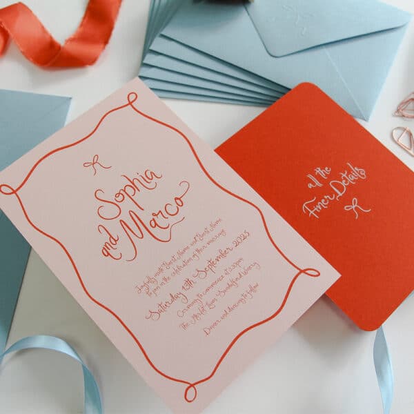A Wedding Invitation suite including an invitation with blush pink background and orange printing. The design uses organic flowing fonts and hand drawn bows and boarders. There are blue envelopes and satin ribbon in the background.